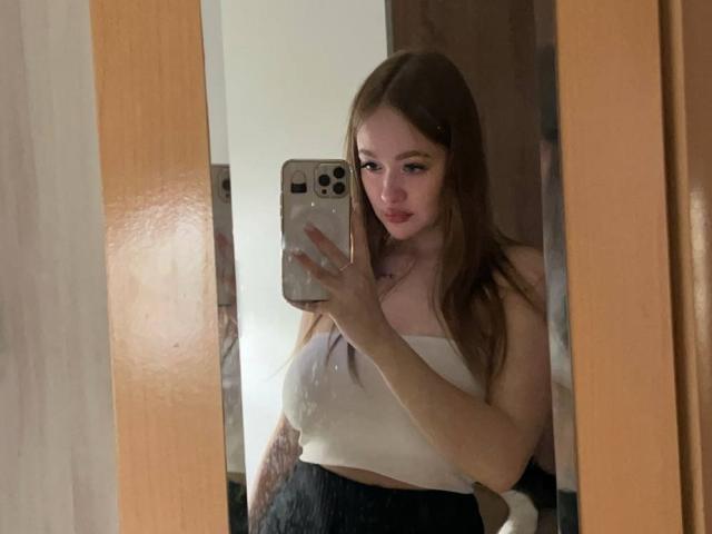 Selfie de PamulaMetzner, cabelo castanho comprido, blusa branca sem alças, saia preta, segurando o celular em frente ao espelho, sexy. expressão, leve sombra no lado esquerdo, moldura de madeira no lado direito, luz natural.