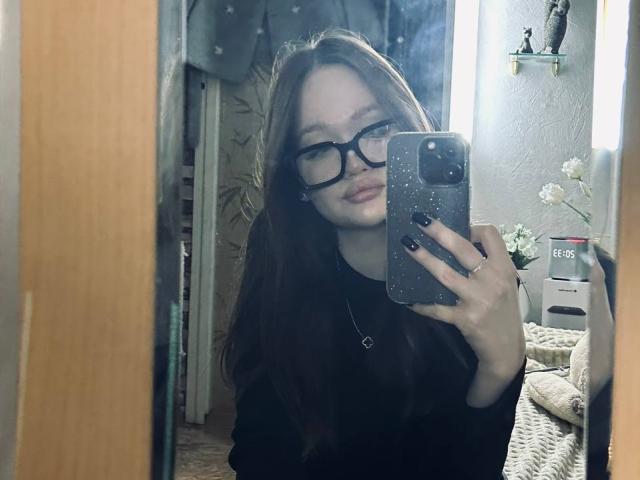 PamulaMetzner, uma mulher magra com cabelos castanhos longos e óculos pretos, tira uma selfie em frente ao espelho com um celular com manchas. Ela está usando uma blusa preta, com um colar visível. Incrivelmente sexy..