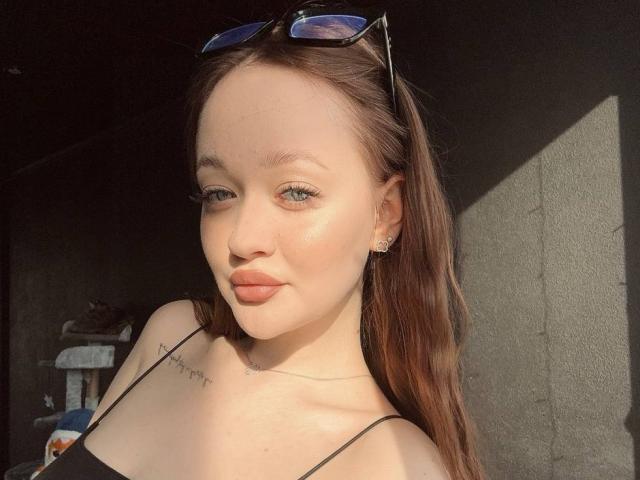 Photograph of PamulaMetzner with light skin, brown hair, and blue eyes, wearing a black strap top, sunglasses on head, sunlight highlighting her face, subtle tattoo on neck, sexy expression.