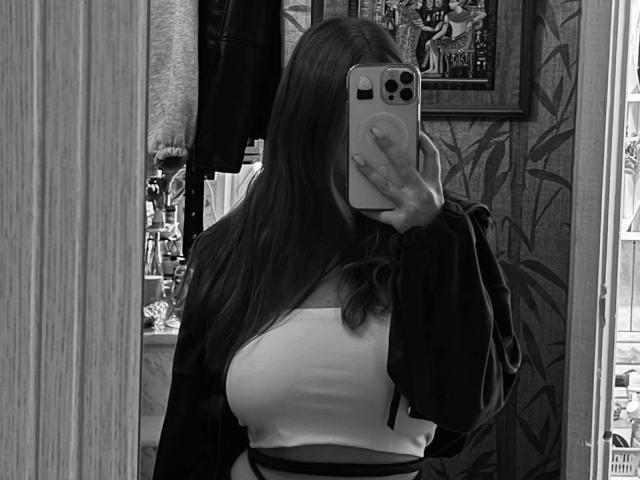 Black and white photo of PamulaMetzner, long hair, holding phone in front of face, large breasts in tight white top, black jacket, standing in room with plants and framed picture. Fucking sexy.