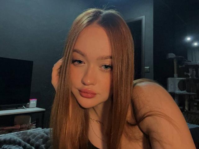 Close-up of PamulaMetzner, a young woman with long, straight brown hair, full lips, and blue eyes, leaning forward with a slight pout. Dimly lit room, black background, soft shadows.