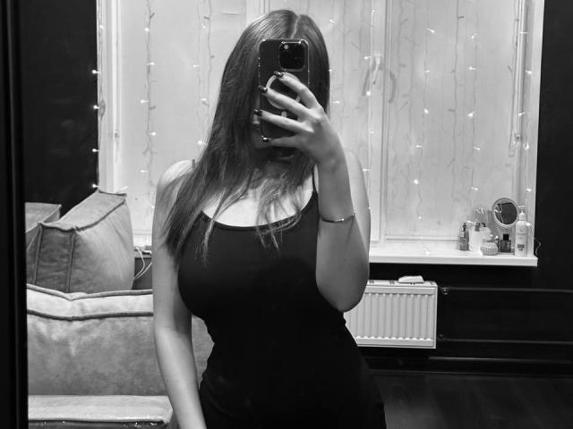 Schwarz-weißes Foto von Pamula Metzner, die ein Telefon vor ihrem Gesicht hält und ein eng anliegendes schwarzes Kleid trägt, wobei sich das Spiegelbild in ein Spiegel. Lichterketten im Hintergrund, Sofa mit Kissen. Finger auf dem Handy, Arm leicht angehoben..