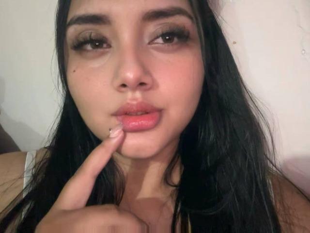 Close-up of AmaliaDalton, a young woman with long black hair, fair skin, and dark eyes, biting her pink finger while gazing seductively at the camera. Glossy lips, heavy eyelids.