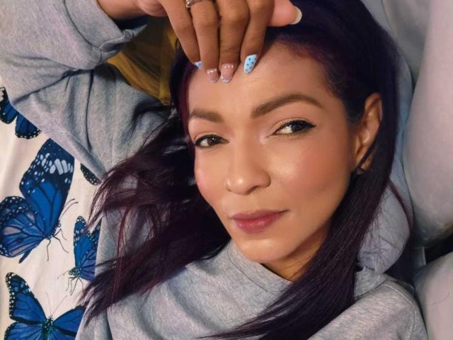 Close-up van Hanna, liggend, hand op haar voorhoofd, draagt een grijze sweater, achtergrond met een blauw patroon van vlinders. lang donker haar, subtiele make-up, lichte glimlach, blauwe nagellak, uitdagende uitstraling..