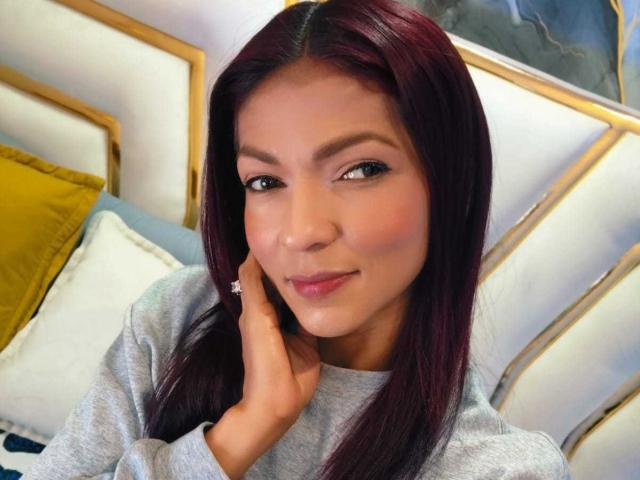 Fotografía de primer plano de HannaPretty, una atractiva mujer asiática con cabello largo y negro, que lleva un suéter gris y tiene una mano tocándose la mejilla. Sonrisa suave, ligero rubor, cama con almohada amarilla en el fondo..