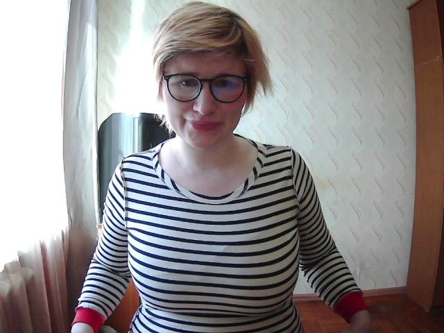 Sabrina, avec ses courts cheveux blonds, portant une chemise rayée noir et blanc, des bracelets rouges et des lunettes, est penchée. Légèrement de face, regard direct, lumière du soleil venant de la gauche, mur texturé en arrière-plan, légère ombre sur la droite..