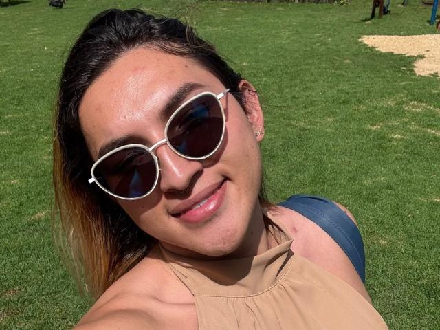 TommyTS with medium skin tone, dark brown hair, and sunglasses, smiling at the camera, wearing a beige top. Green grass in the background. Sunlight highlights TommyTS's face. Fucking hot.