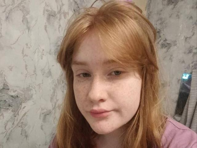 Gros plan de SheilahSexy, avec des cheveux roux, des taches de rousseur et une peau claire, portant une chemise violette, regardant directement l'appareil photo. avec une expression légèrement séduisante. Fond avec une texture de marbre. Tellement mignon..