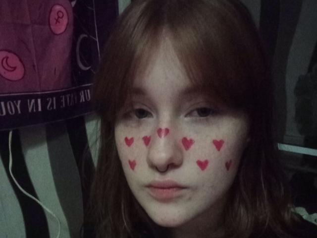 Fotografía de primer plano de SheilahSexy con piel pálida, cabello castaño y marcas en forma de corazón rojas en sus mejillas. Ella tiene una expresión neutra, sentada frente a un fondo oscuro con un diseño..