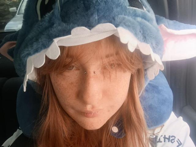 Close-up de SheilahSexy com cabelo ruivo, sardas e um chapéu azul em formato de tubarão com babados brancos. Ela está inclinada para frente, com os olhos semicerrados, e a luz do sol realça seu rosto e chapéu..