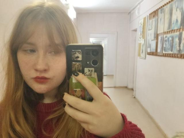 Selfie of SheilahSexy with red lipstick, freckles, and long brown hair, holding a phone with a sticker case, wearing a red sweater, standing in a gallery hallway with framed pictures.