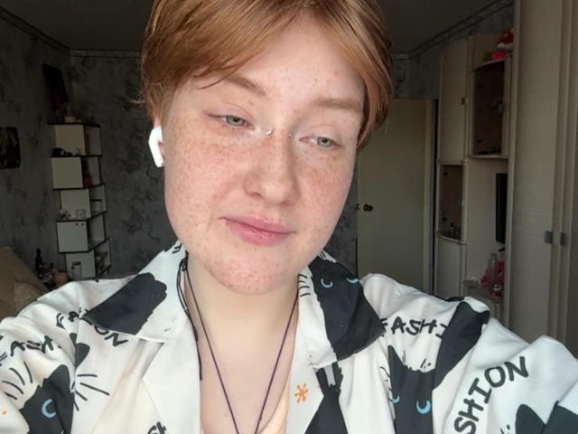 Close-up van SheilahSexy, een rodeharige vrouw met sproeten en kort haar, die een zwart-wit geruit shirt draagt en witte oordopjes heeft. en een ketting, kijkend in de camera met een lichte glimlach. Achtergrond van de kamer.