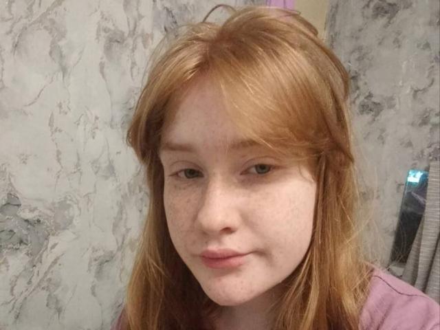 Nahaufnahme von SheilahSexy mit heller Haut, roten Haaren und Sommersprossen, die ein lila Hemd trägt. Ihr Gesichtsausdruck ist neutral, und ihre leicht geöffneten Lippen verraten eine spürbare sexuelle Spannung. Marmor-Wand als Hintergrund..