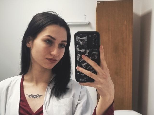 Fotografia de RoxxanneBoby, de pele clara e cabelo preto, vestindo uma túnica branca sobre uma blusa vermelha, segurando um telefone preto. com uma capa floral, refletindo em um espelho, tatuagem no peito.