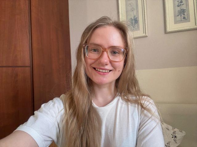 Photographie de missNancy-hot, une jeune femme aux longs cheveux bruns lisses, portant des lunettes orange et un t-shirt blanc. Elle sourit directement à la caméra, assise à l'intérieur, avec des œuvres d'art en bois et encadrées en arrière-plan..
