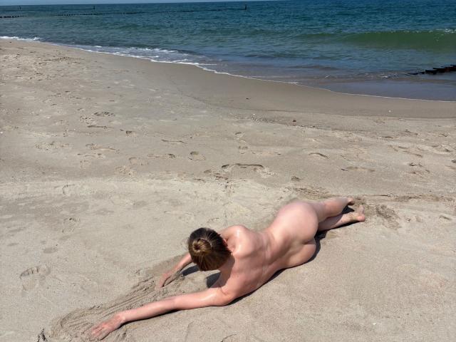 Nude missNancy-hot, lying face down on a sandy beach, arms and legs spread out, back arched, sunbathing, ocean in background, blue waves, wet sand, clear sky, relaxed pose.