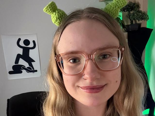 Nahaufnahme von missNancy-hot mit hellbraunen Haaren, die grüne, gestrickte Katzenohren und große, rosa Brillen trägt, lächelnd. mit einer schwarzen Silhouette im Hintergrund. Total süß..