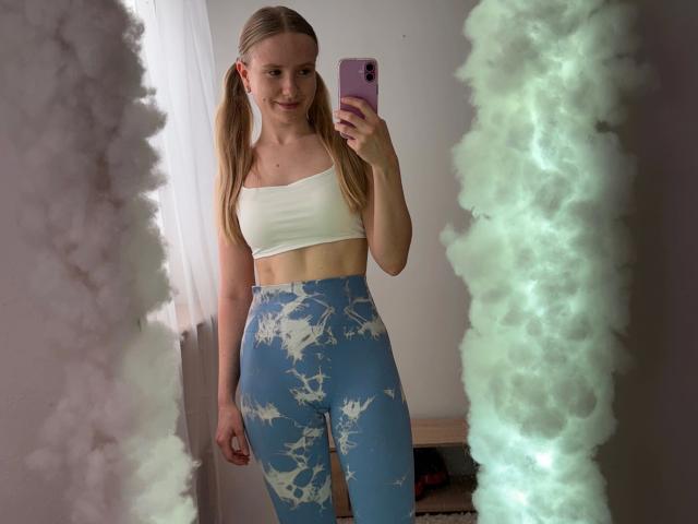 MissNancy, aantrekkelijk met vlechtjes, een witte top en blauwe hoge taille leggings met witte patronen, staat voor gloeiende mist. Met een roze telefoon in de hand, lachend, met zichtbare decolleté en strakke buikspieren..