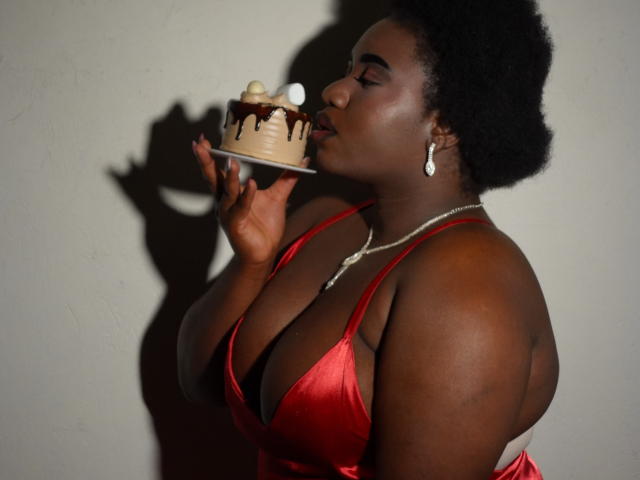 Photograph of a dark-skinned woman, Dilay, with a curvy physique and large breasts, wearing a red satin halter top, holding a dessert close to her lips. Shadows cast on the wall.