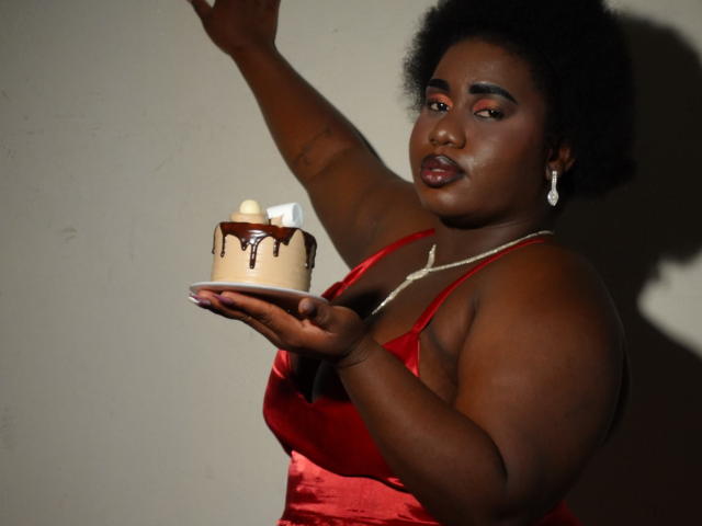 Fotografía de una mujer de piel oscura, con una figura completa y curvilínea y senos grandes, vestida con un vestido rojo y perlas. collar, sosteniendo un pastel encendido con chocolate goteando. Su brazo está levantado..