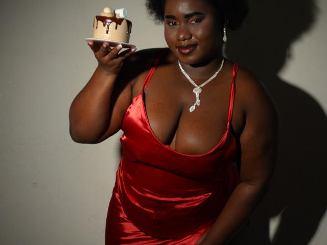 Fotografie einer dunkelhäutigen Frau, Dilay, in einem roten Seidenkleid, die eine angezündete Geburtstagstorte hält. Sie hat große Brüste, trägt eine Halskette und es ist ein Schatten an der Wand zu sehen. Verdammt heiß..