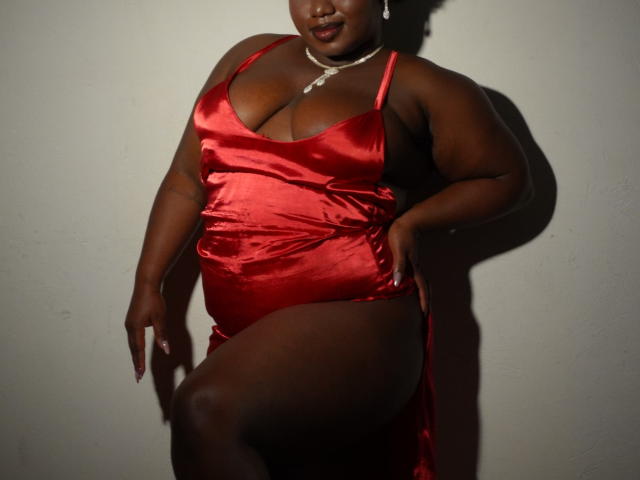 Dilay, una mujer de piel oscura con un cuerpo curvilíneo y de talla grande, lleva un vestido rojo de satén brillante con un escote pronunciado. Ella está de pie frente a una pared lisa, levantando su vestido. Las sombras resaltan sus curvas..