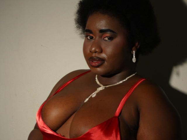 Fotografia di Dilay, una donna nera formosa con grandi seni, vestita con un abito rosso in raso scollato, che indossa una collana di perle e orecchini, appoggiati contro un muro bianco, ombre sul viso.