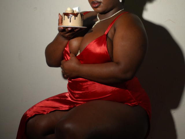 Dilay with dark skin, wearing a red satin dress, holding a lit candle, shadows highlighting her curves and cleavage. Pearl necklace, lips slightly parted, intense light, erotic tension.