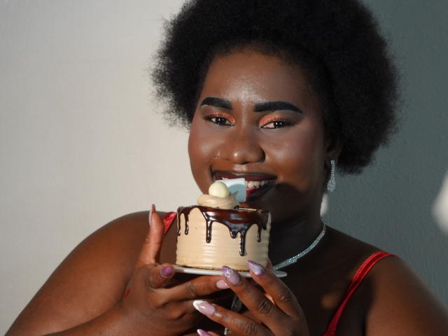 Dilay, avec sa peau foncée et ses cheveux afro naturels, porte un haut rouge et tient un cupcake trempé dans du chocolat, avec du glaçage et de la crème fouettée. crème, qu'elle léchait de manière suggestive, ongles violets, éclairage vif, tension sexuelle.