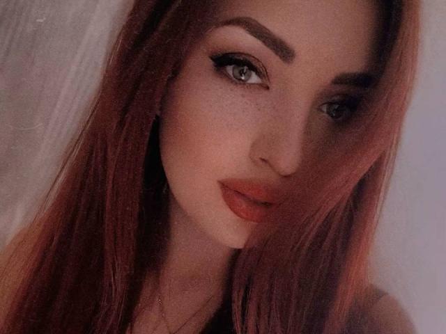 Primer plano de FireeFely, una joven con cabello largo y rojo, delineador de ojos negro intenso y lápiz labial rojo. Su mirada seductora y su ligera sonrisa crean una tensión intensa y sexual. La luz suave resalta su rostro..