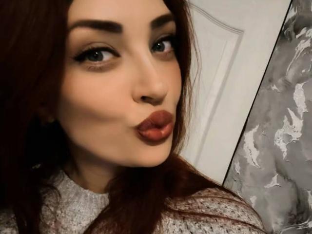 Nahaufnahme von FireeFely, einer attraktiven Frau mit dicken, braunen Haaren und vollen Lippen, die zu einem Kuss geformt sind, die ein graues, strukturiertes Oberteil trägt. Ihr intensiver Blick und ihr sinnlicher Ausdruck strahlen eine sexuelle Spannung aus..