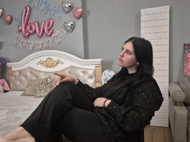 Esfil, com cabelo preto, vestindo um vestido preto, está sentada em uma cama com cabeceira estofada, com as pernas cruzadas, olhando para... câmera, cercada por decorações de corações rosa e prateados.