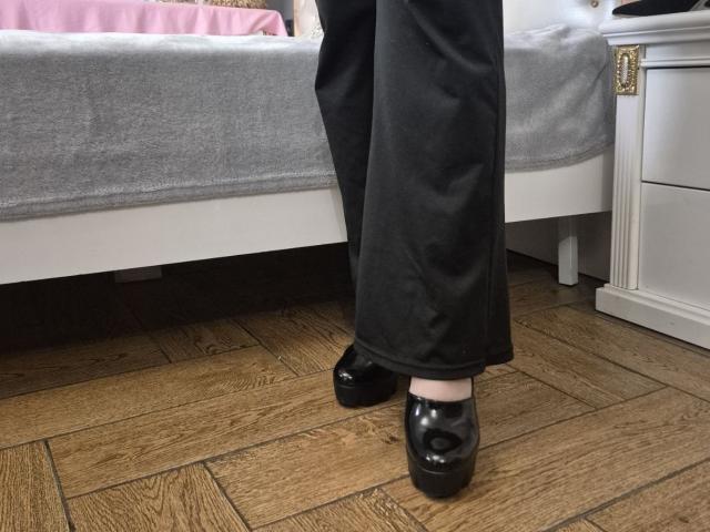 As calças pretas e os sapatos pretos brilhantes de Esfil estão em um chão de madeira, ao lado de uma cama com um cobertor cinza. A pele clara de Esfil é visível logo acima dos sapatos. Que demais..
