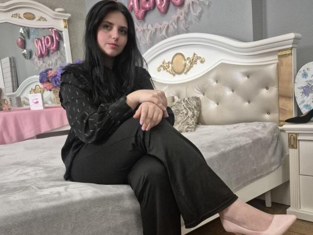 Esfil, con lunghi capelli neri, seduta su un letto bianco imbottito, indossa una maglietta nera a pois e pantaloni neri, scarpe rosa, aspetto sexy. atmosfera, il letto ha una testiera bianca, decorazioni a forma di cuore rosa..