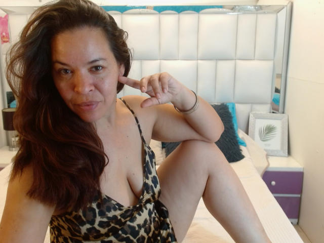 LizethBells, avec de longs cheveux bruns, portant une robe à imprimé léopard à décolleté plongeant, assise sur un lit, pointant son oreille, sexy. ambiance, décolleté, chambre avec tête de lit blanche, éclairage doux.