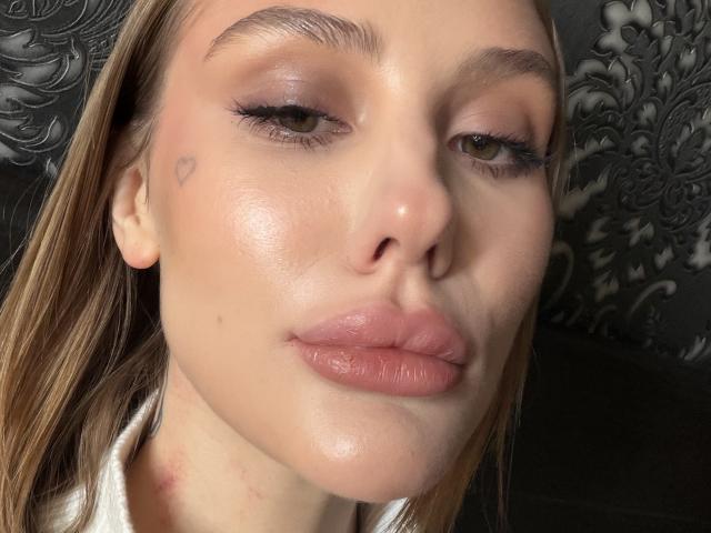 Close-up of SensualMila with shiny skin, full lips, and a small heart tattoo on her cheek. She has half-lidded eyes and a subtle smirk. Black floral pattern in the background.