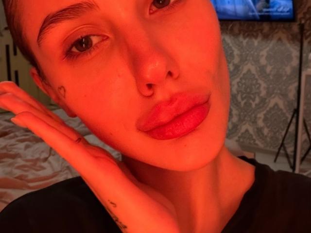 Close-up foto van SensualMila, met een gloeiende rode gloed, kort bruin haar, een opvallende neusring, volle rode lippen, hand die iets aanraakt. Gezicht, zwart shirt, tatoeage op de hand, blauw televisiescherm op de achtergrond..