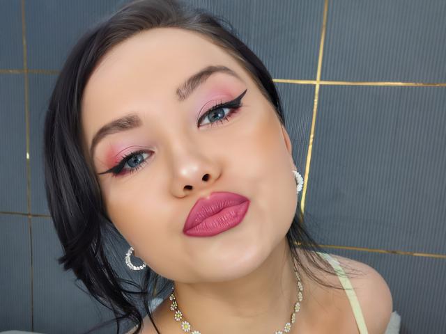Fotografía de primer plano de KimHonney, de piel clara y cabello negro, con sombra de ojos rosa intensa, delineador de ojos negro grueso, lápiz labial rojo. aretes de aro dorados, collar, mirando ligeramente hacia arriba, tensión sexual, rostro absolutamente hermoso..