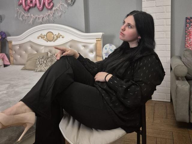 Esfil, con i capelli neri e la pelle chiara, è seduta su una sedia, con le gambe incrociate, indossa una maglietta e pantaloni a pois neri e tacchi beige. Letto lussuoso con testiera bianca e insegna rosa con la scritta "LOVE" sullo sfondo..