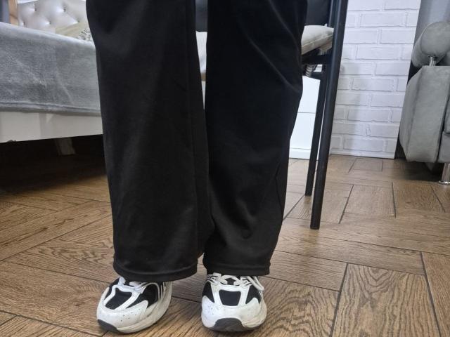 Le pantalon noir d'Esfil met en valeur ses baskets blanches et noires sur un plancher en bois. Un canapé gris et un mur en briques blanches se trouvent en arrière-plan. Esfil se tient debout, légèrement penché..