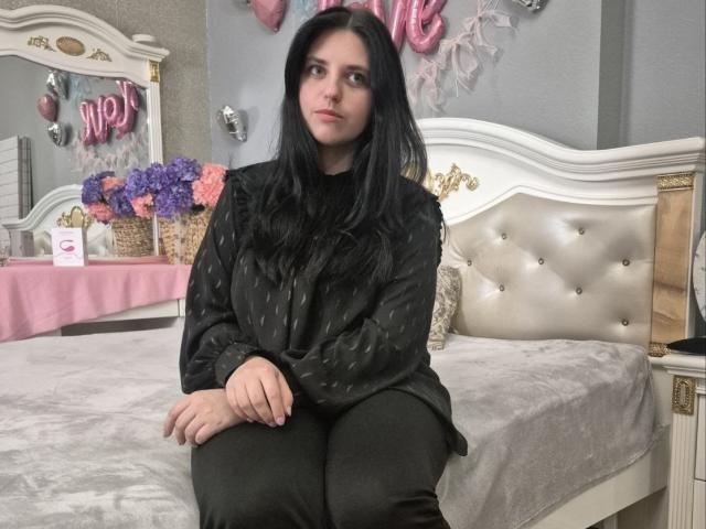 Esfil mit langem, schwarzem Haar, sitzt auf einem weißen, gepolsterten Bett und trägt ein schwarzes, gepunktetes Hemd und schwarze Hosen. Rosa Ballons und eine Blumenarrangements im Hintergrund. Verführerischer Blick..