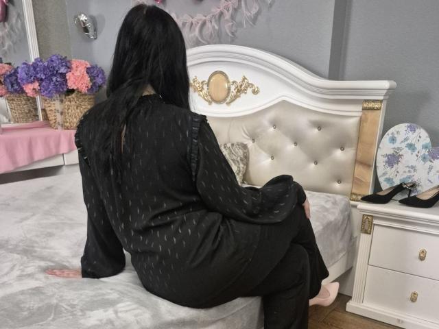 Esfil, met lang zwart haar, zittend op een wit bed met een bekleding van veertjes, met de rug naar de camera gericht, gekleed in een zwarte badjas met stippen, benen licht gespreid, rechterhand rustend op het bed, sensuele spanning.