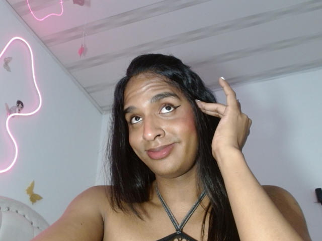 Fotografie von IdiiaLatina mit mittelbrauner Haut, schwarzem Haar und dezentem Make-up, die lächelnd ihre Haare berührt. Eine Hand, die ein schwarzes Top mit Trägern trägt, mit einer neonrosa Umrandung im Hintergrund..