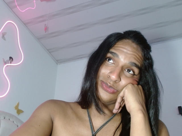 Fotografie von IdiiaLatina, einer nackten Frau mit langen schwarzen Haaren, brauner Haut und vollen Lippen, die auf ihrer Hand ruht und in die Ferne blickt. verführerisch mit einem leichten Lächeln, Neonlichter im Hintergrund..