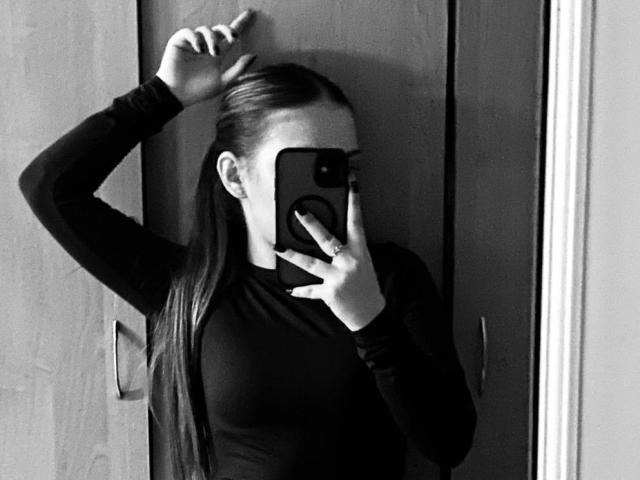 Black and white photograph of ZoieHurt, standing against a door, holding a phone over her face with her right hand, left hand raised above her head. Tight, dark shirt. Shadowed, moody lighting.