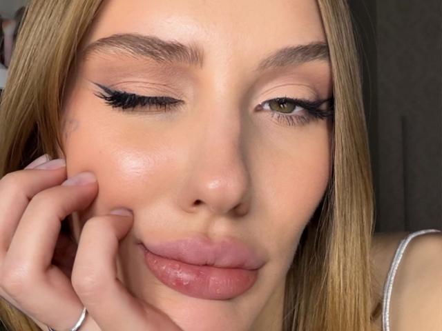 Close-up photo of SensualMila, winking with one eye, full lips slightly parted, hand touching cheek, long blonde hair, thick black eyeliner, natural skin texture, wearing a white spaghetti strap.