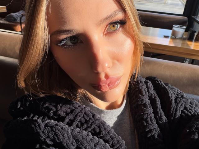 Close-up photo of SensualMila, sunlight illuminating her face, brown eyes, and blonde hair. She's wearing a black, chunky-knit sweater, with shadows creating a sensual, intense look. Her lips are slightly parted.