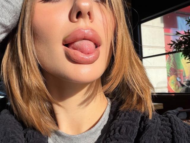 Close-up of SensualMila with her tongue sticking out, glossy pink lips, light brown shoulder-length hair, wearing a gray top and black jacket. Sunlight highlights her facial features, background blurred.