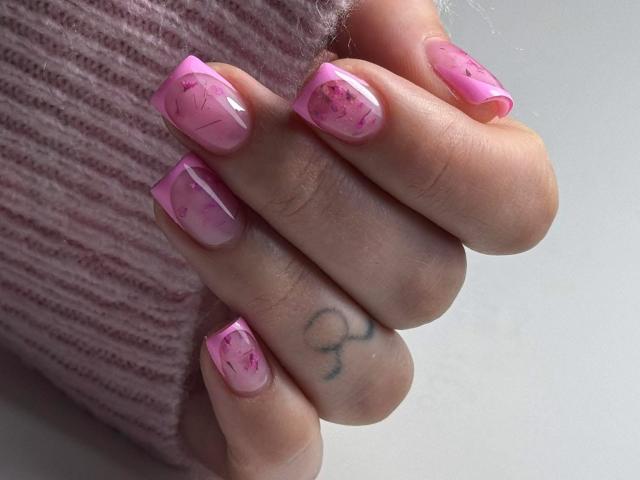 Primo piano della mano di SarahFontain, con unghie rosa scintillanti, un piccolo tatuaggio nero sull'anulare, che indossa un anello rosa con una texture particolare. maglione. Le unghie brillano alla luce, la mano è parzialmente serrata..