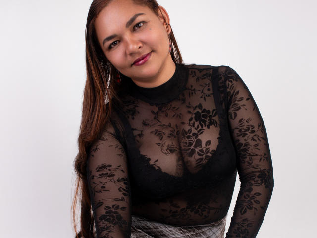 VivianBig mit langem, braunem Haar, trägt eine durchsichtige, schwarze Spitzenbluse, die ihre Brustwarzen freilegt, und lehnt sich leicht nach vorne. Lächeln. Weißer Hintergrund. Sexuelle Spannung durch ihren selbstbewussten Blick..